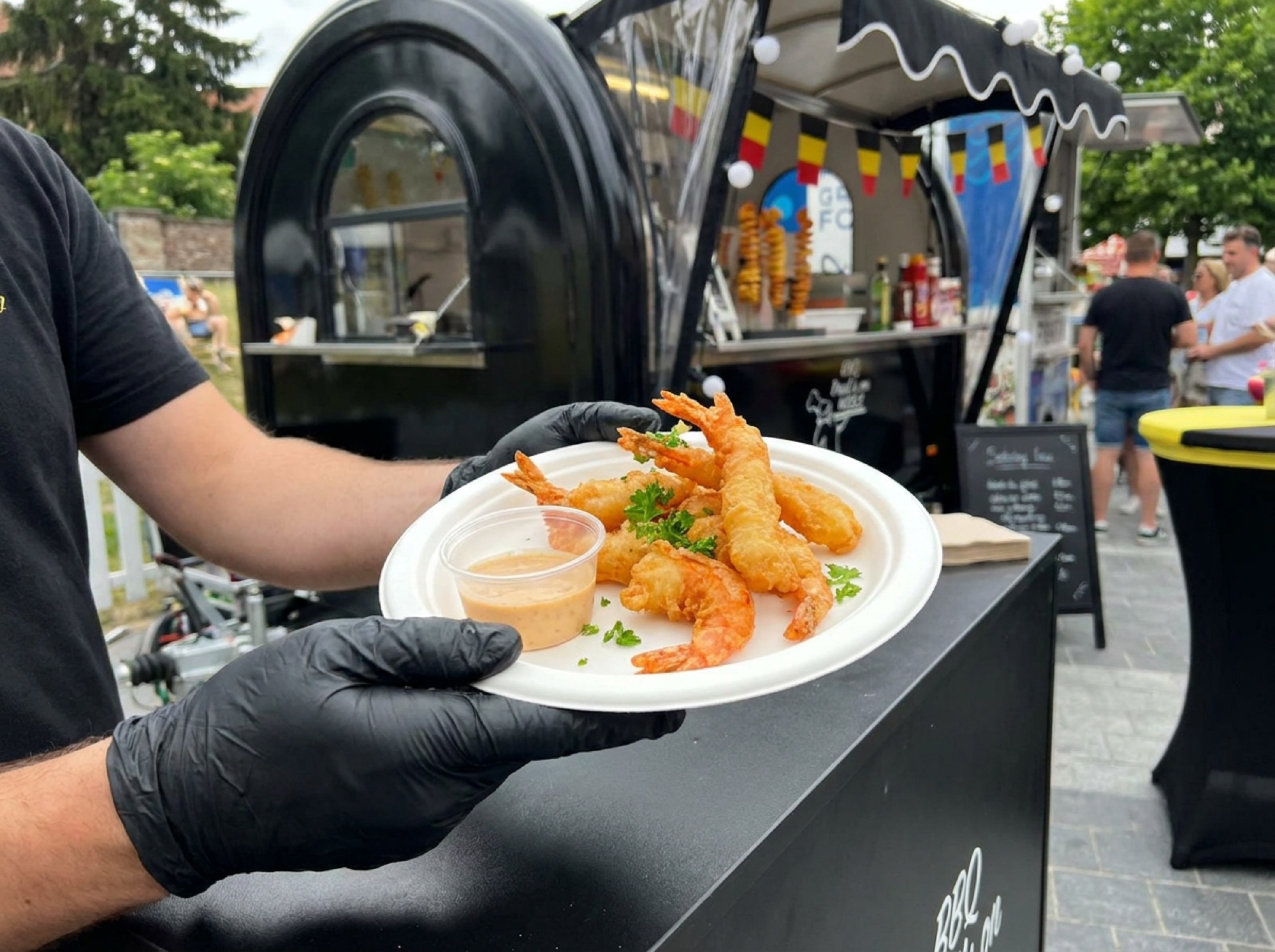 Tempura tijgergarnalen op wit suikerrietbord met dipsaus, vastgehouden met zwarte handschoenen
